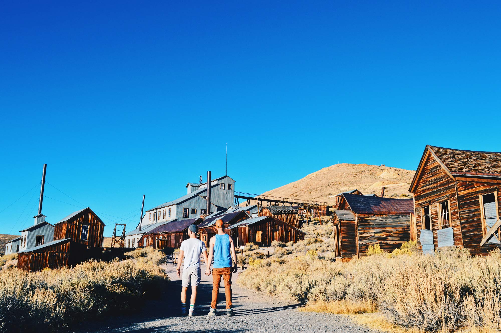 Gay Couple Travelers hand in hand in front Standard Consolidated Mining Company Stamp Mill | Ghost Town Bodie State Historic Park California © CoupleofMen.com