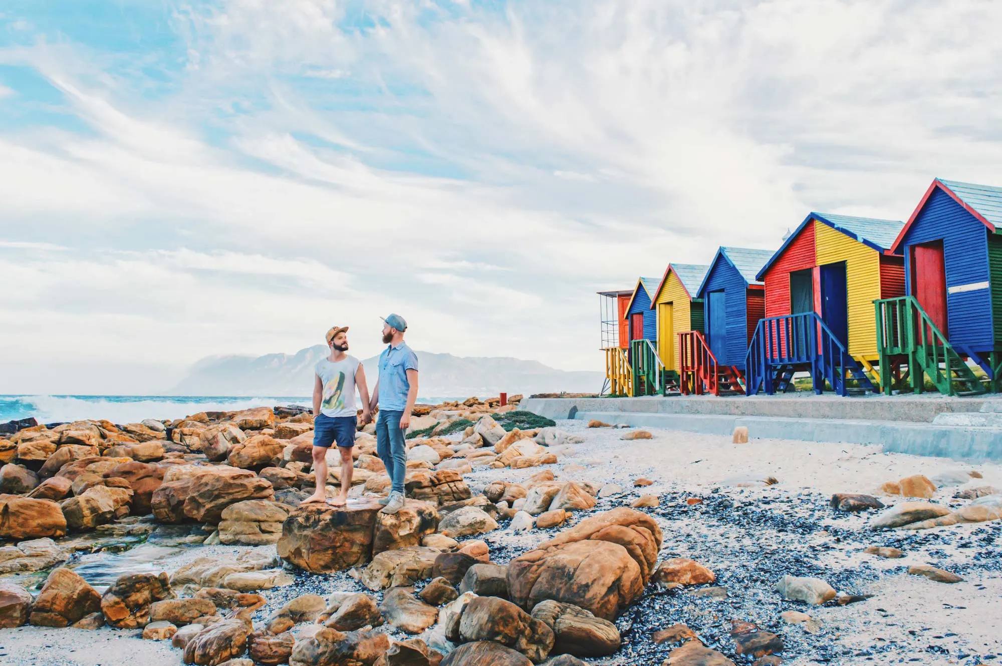 Gay Couple City Trip Cape Town Gay Travel Cape Town: Adventure Days of a Gay Couple standing hand-in-hand in front of colored beach houses in the South of the Mother City © Coupleofmen.com