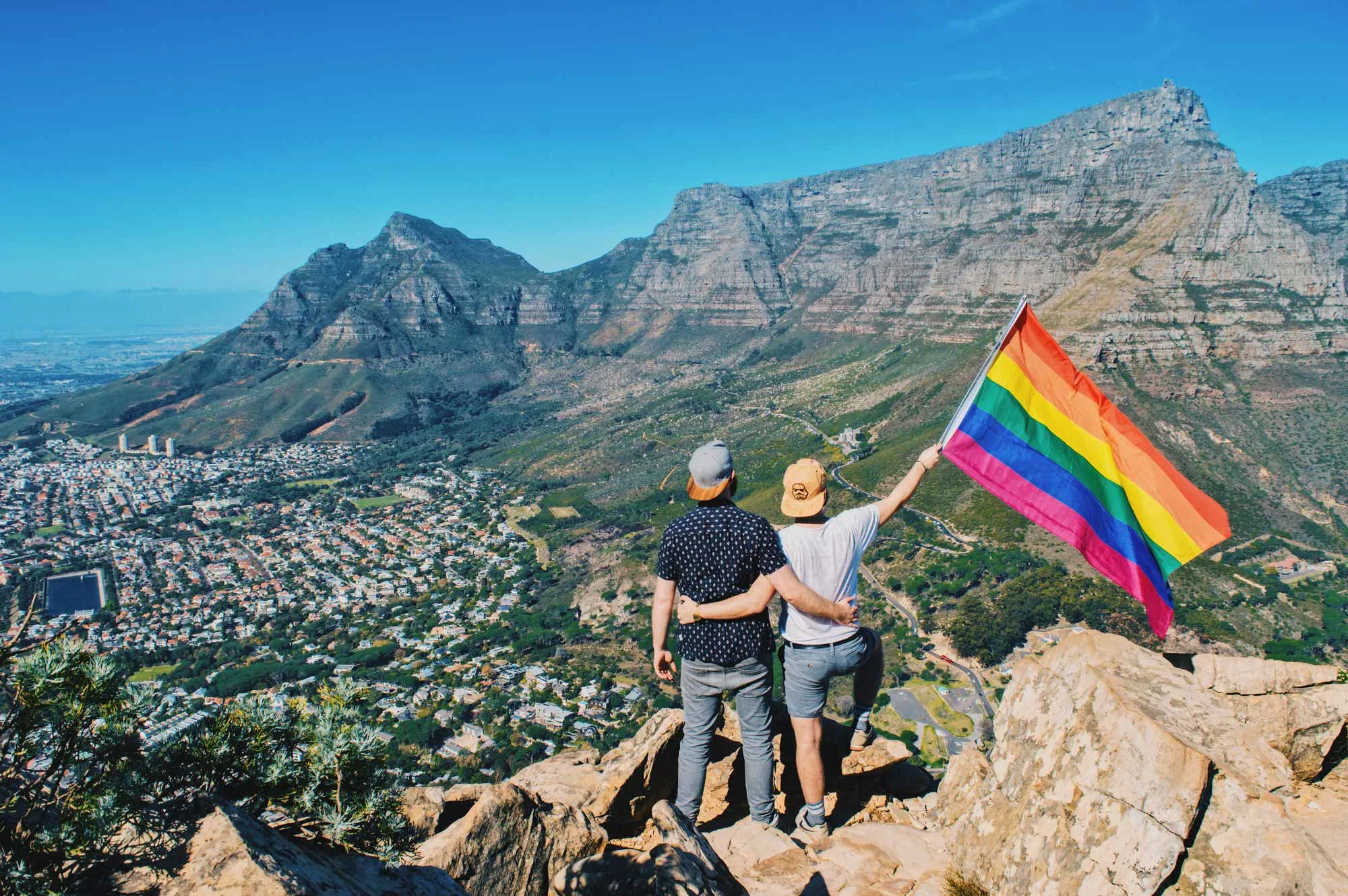 Gay Couple standing hand-in-hand on Lion's Head with a rainbow flag overlooking Cape Town & Tabel Mountain © Coupleofmen.com