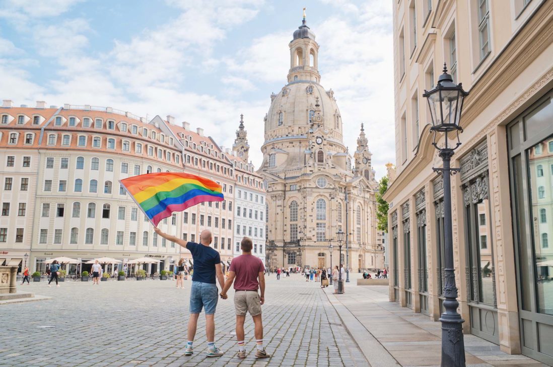 Dresden Gay City Trip: Gay Couple waving a rainbow flag in front of the Church of Our Lady (German: Frauenkirche) © Coupleofmen.com