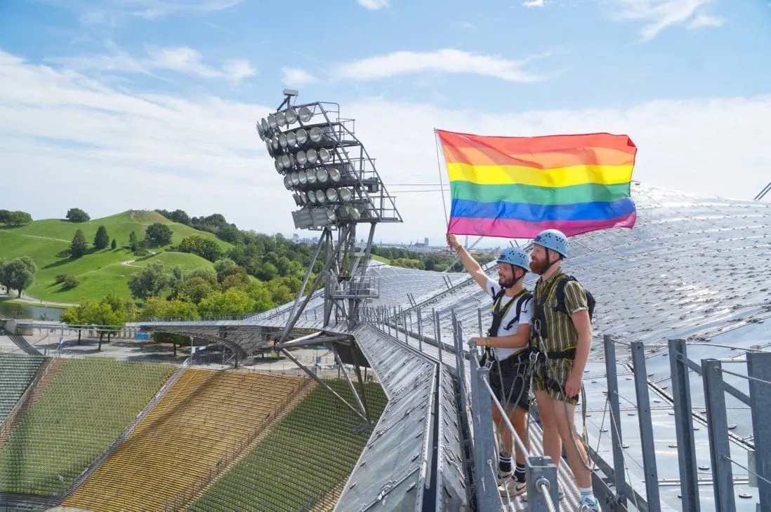 Munich Gay City Trip Proudly waving the rainbow flag as a gay couple on the rooftop of the Olympic Stadium from 1972 © Coupleofmen.com