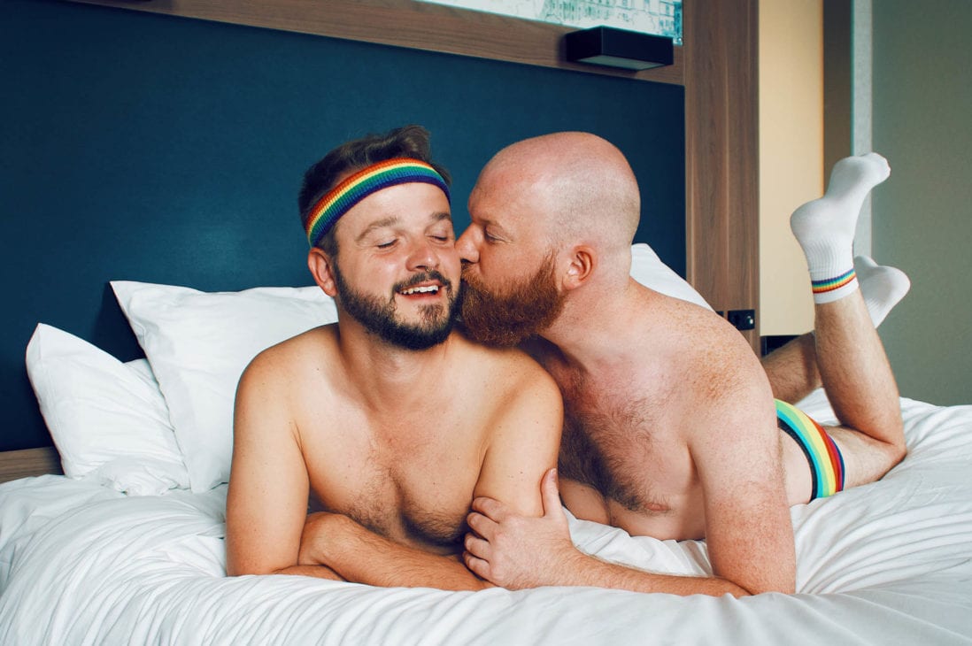 Wearing the rainbow pride gear even in their hotel bed: Gay Couple Travel Blogger laying arm-in-arm in the LGBTQ+ friendly design Hotel in Munich, Germany © Coupleofmen.com