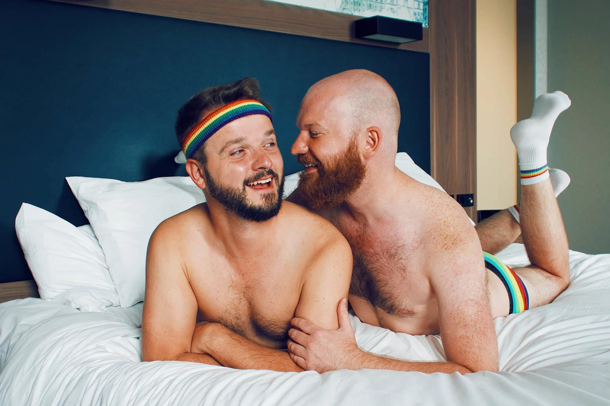 Wearing the rainbow pride gear even in their hotel bed: Gay Couple Travel Blogger laying arm-in-arm in the gay-friendly design Hotel Aloft in Munich, Germany © Coupleofmen.com