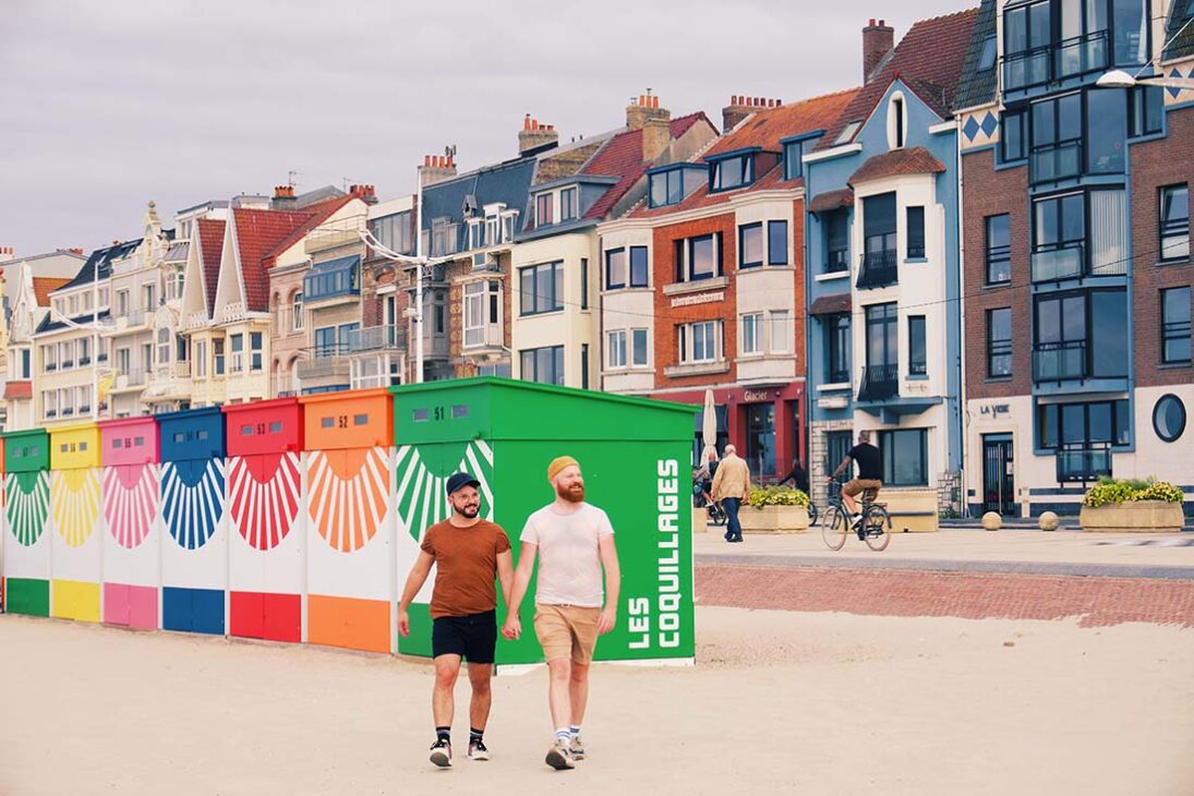 Hand-hand-hand walking along the beach of Dunkerque in front of the colorful beach houses © Coupleofmen.com