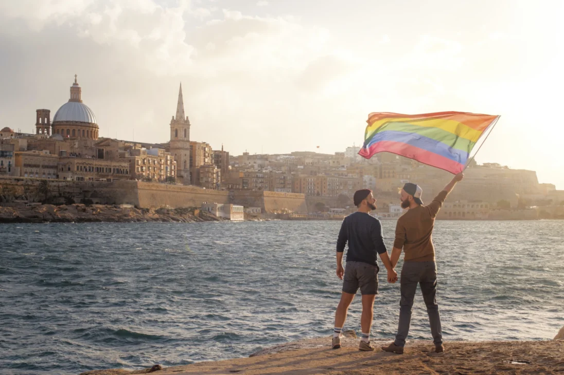 A gay couple of men holding hands and waving a rainbow flag in front of the city of Valletta © coupleofmen.com