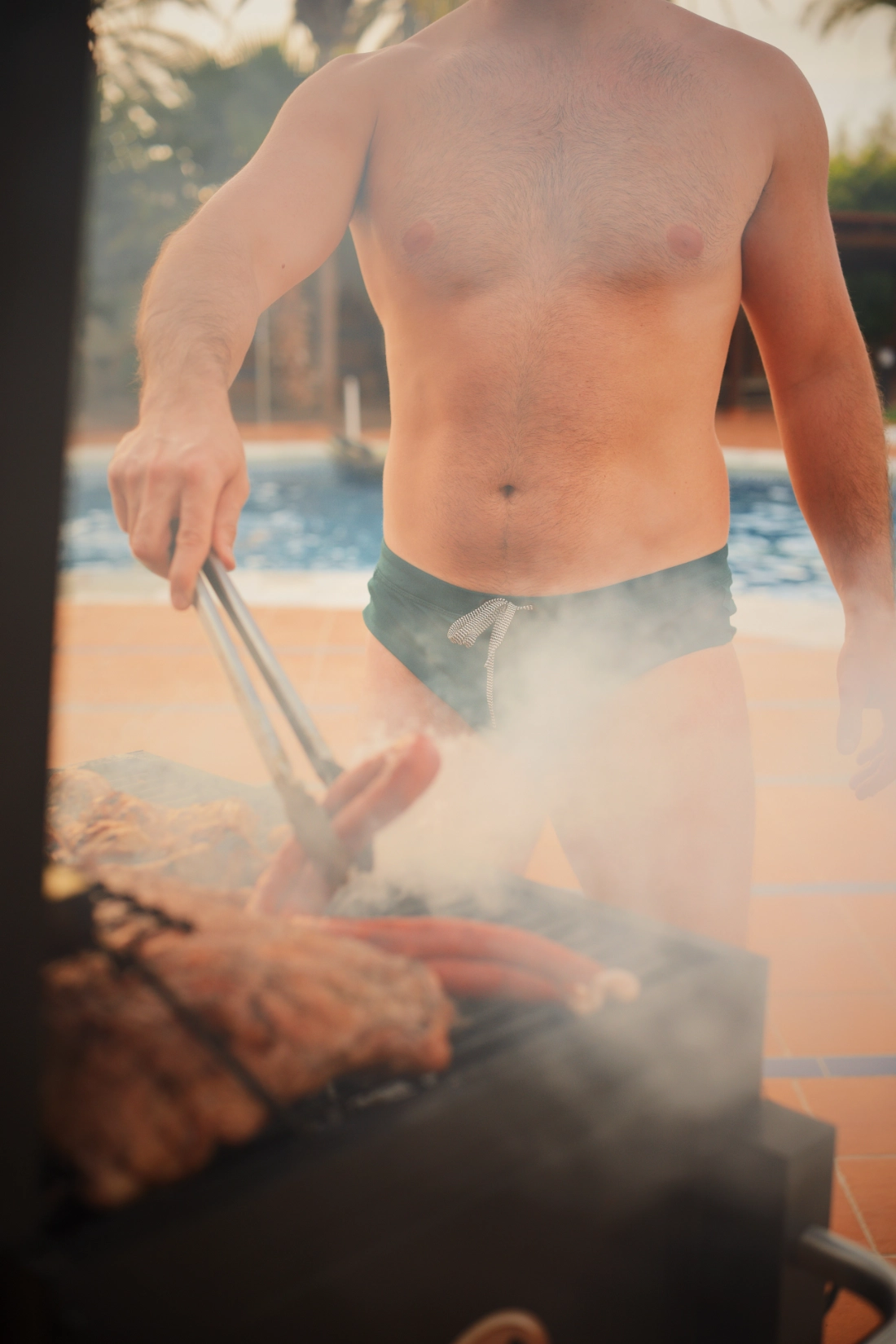 A sexy men is barbecuing while only wearing his speedo.