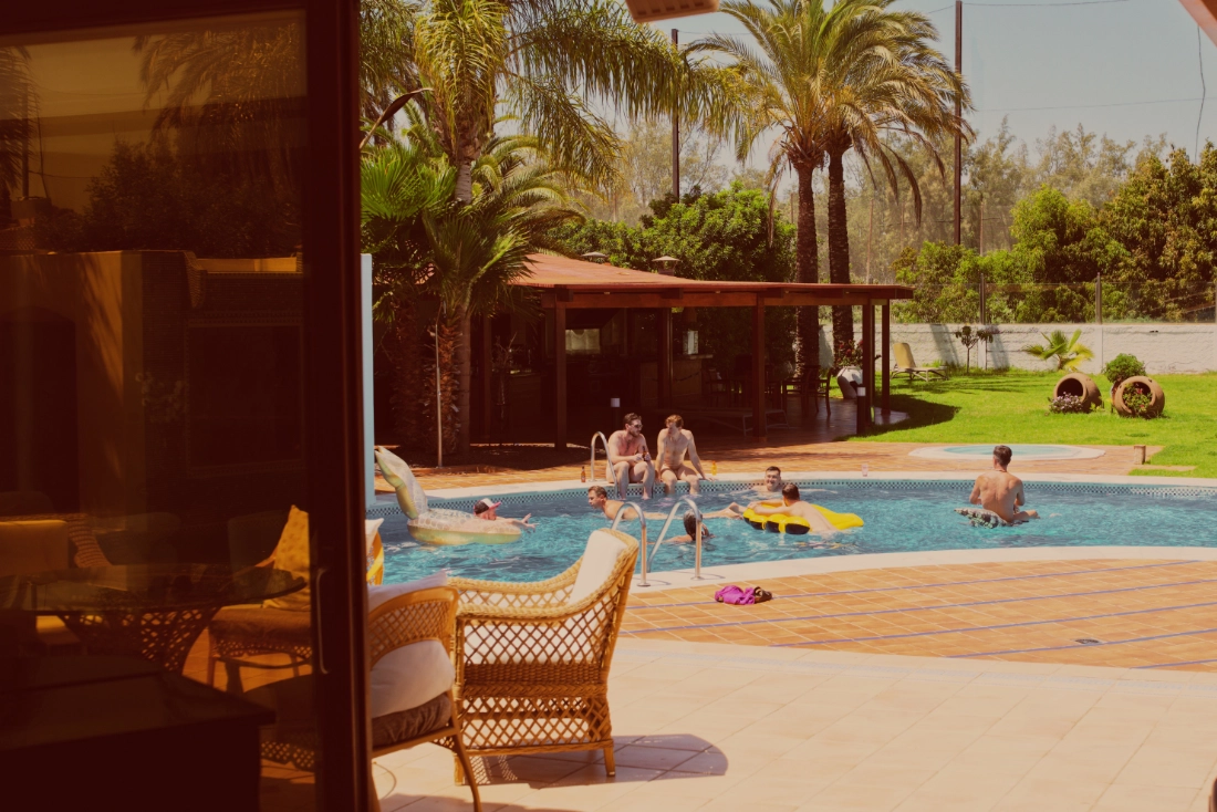 a pool with eight naked men chilling in the water or on the sides while talking and drinking and enjoying the sun.