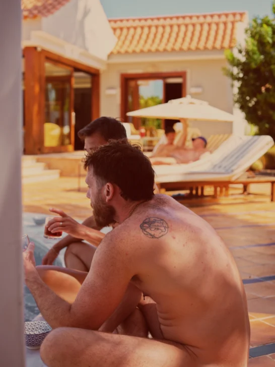 Two men sitting naked at the pool while holding a drink and talking while two othe rmen are talking while laying naked underneath a parasol in front of the Gran Canaria Villa. Clothing-optional Gay Vacation Gran Canaria