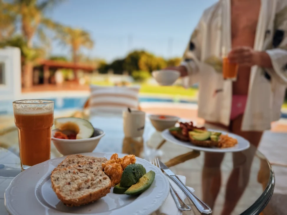 A plate with a breakfast of bread, avocado and scrambled eggs next to a glass of fresh juice and a small bowl of fruits standing on a glass table while a men in speedo's is putting down his drink. In the back the Naked Villa pool and garden.