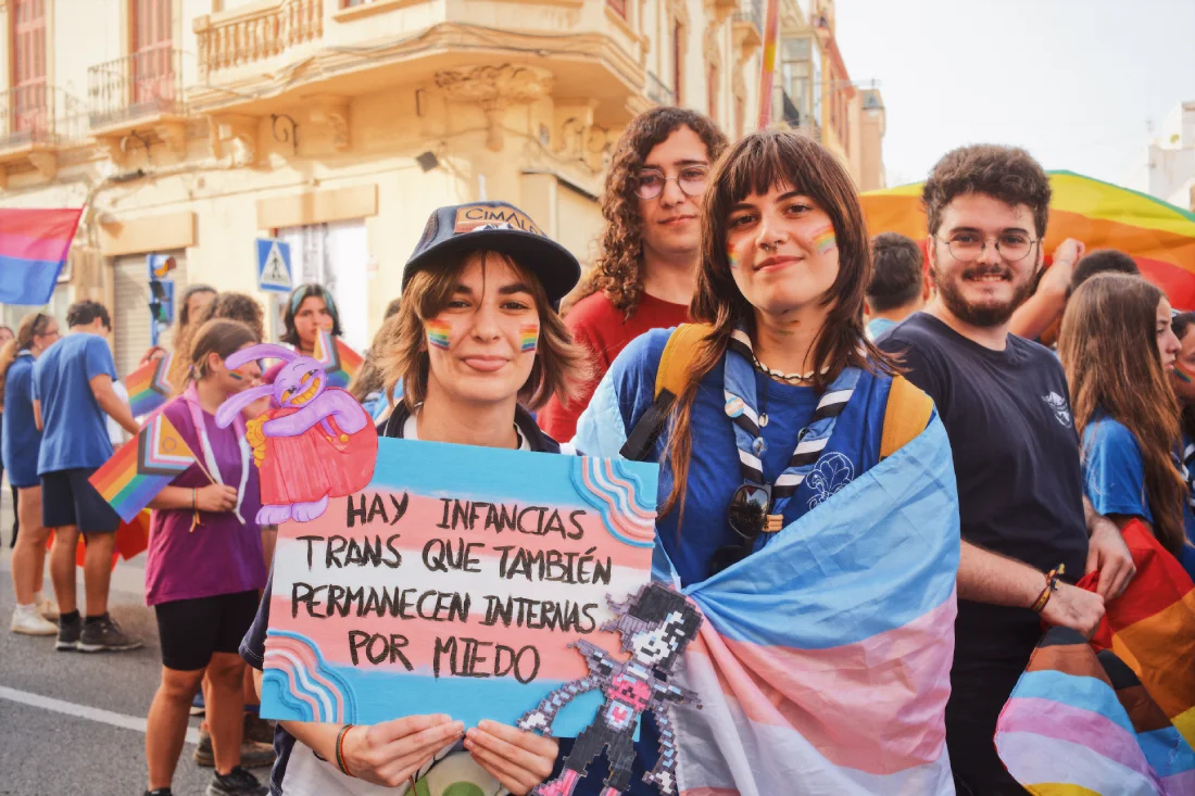 Young Pride participants in Melilla holding a colorful sign reading &ldquo;Hay infancias trans que tambi&eacute;n permanecen internas por miedo&rdquo; and wearing trans and rainbow flags at North Africa Pride